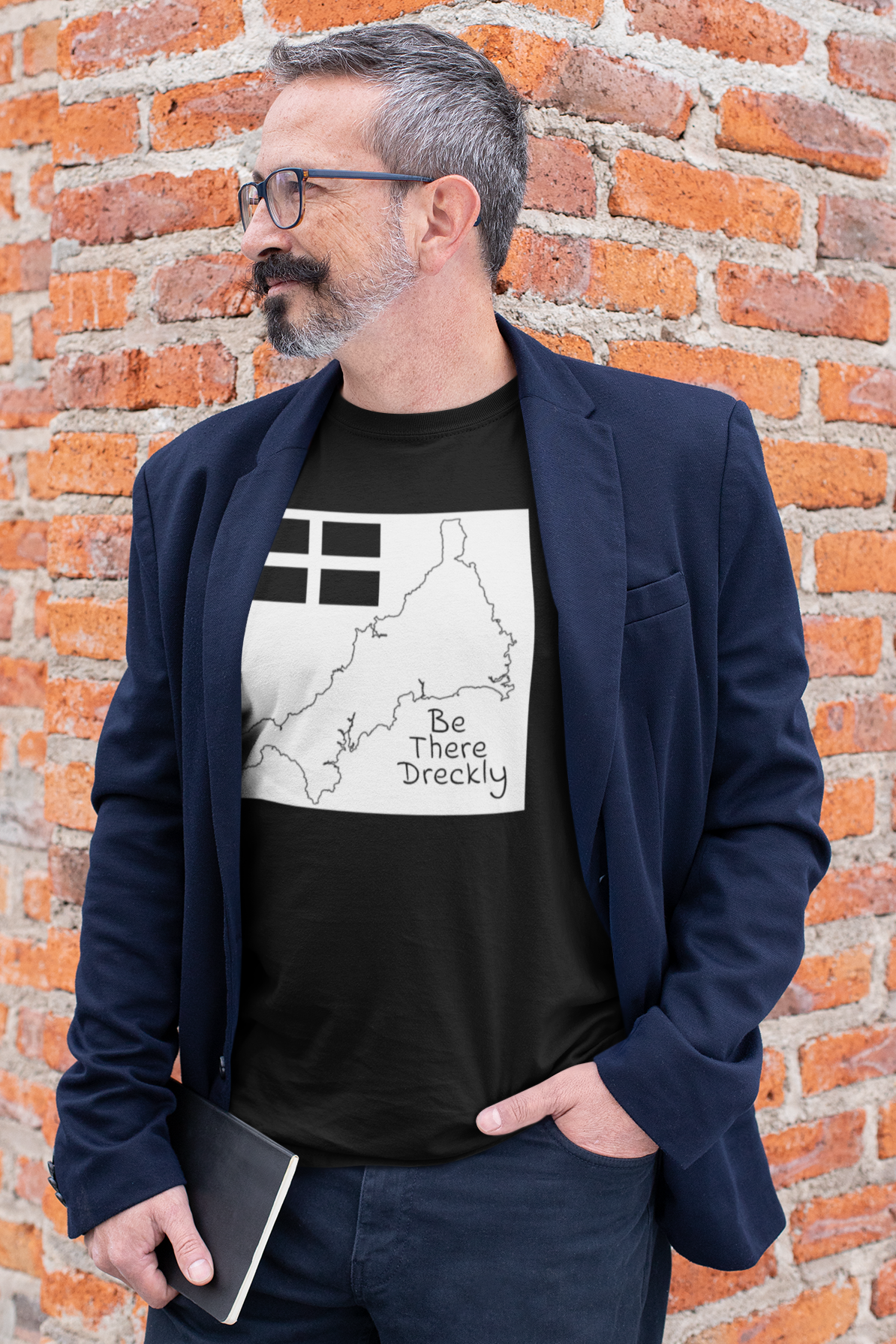 Man wearing black Be There Dreckly Cornwall t-shirt with navy blazer, brick wall background