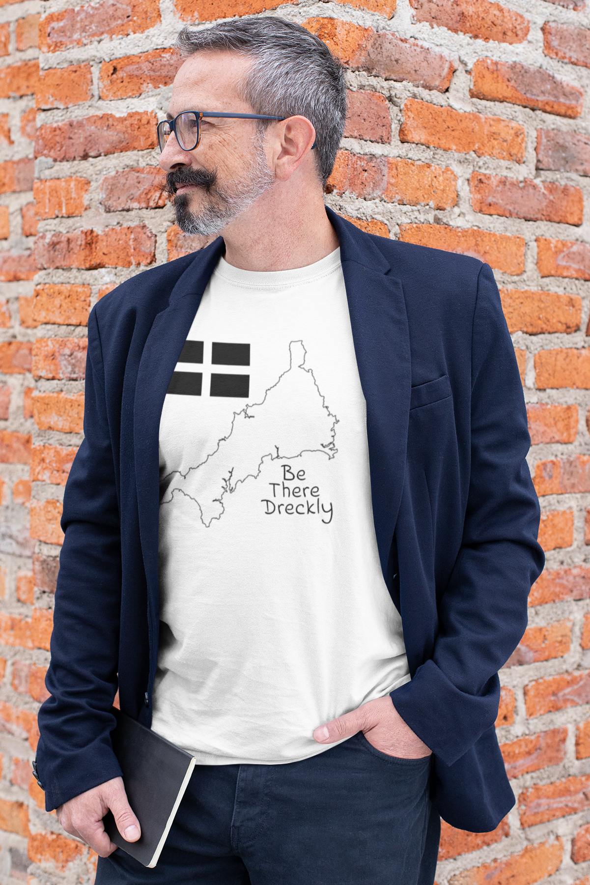Man wearing white Be There Dreckly Cornwall t-shirt with navy blazer, brick wall background