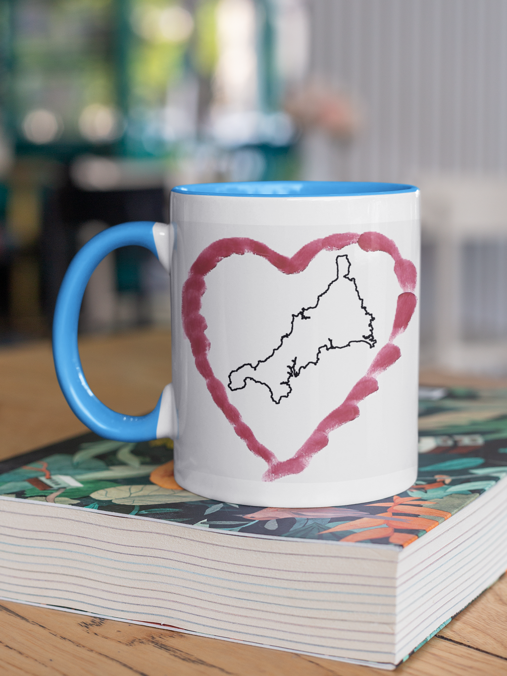 I Love Cornwall heart map mug with light blue handle on patterned book, Cornwall outline design