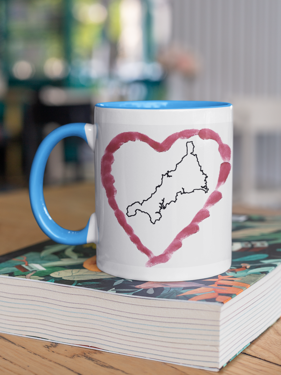 I Love Cornwall heart map mug with light blue handle on patterned book, Cornwall outline design
