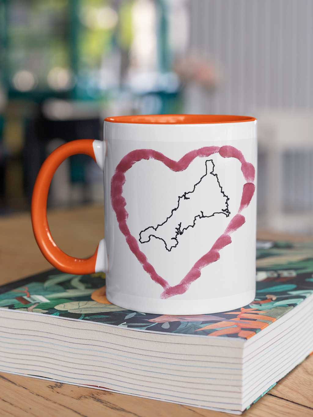 I Love Cornwall heart map mug with orange handle on patterned book, Cornwall outline design