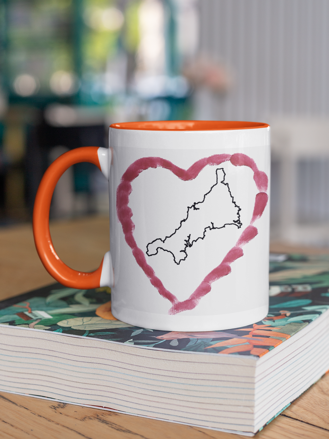 I Love Cornwall heart map mug with orange handle on patterned book, Cornwall outline design