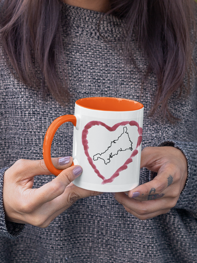 Hands holding I Love Cornwall heart map mug with orange handle, Cornwall outline design