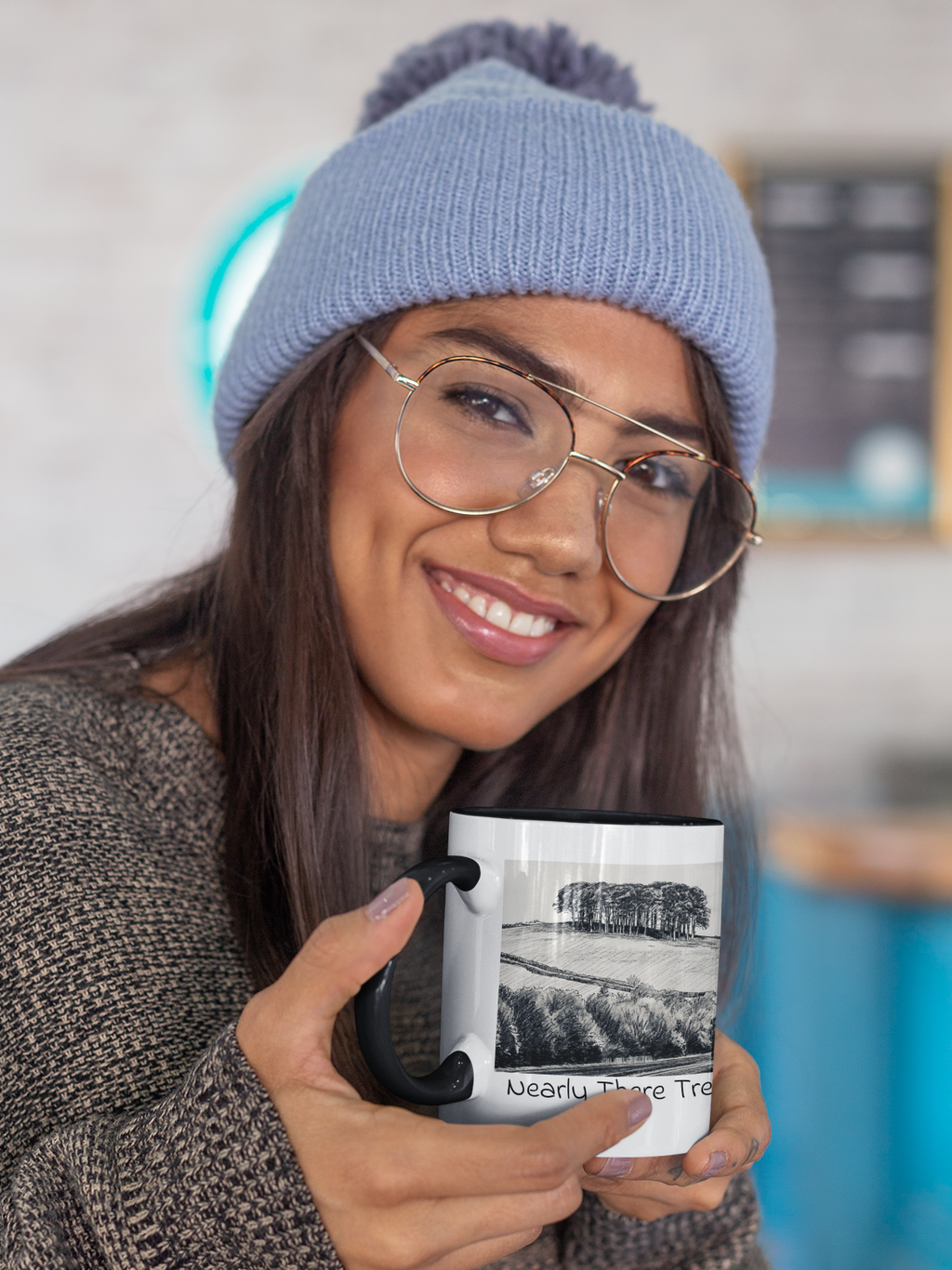 Woman in beanie holding Nearly There Trees Cornwall mug with black handle, tree line pencil sketch design