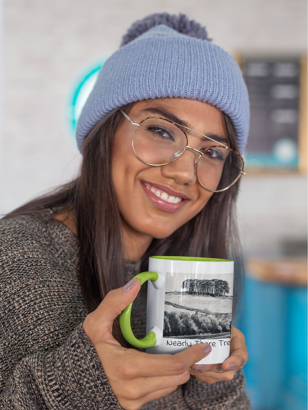 Woman in beanie holding Nearly There Trees mug with green handle, tree line pencil sketch design