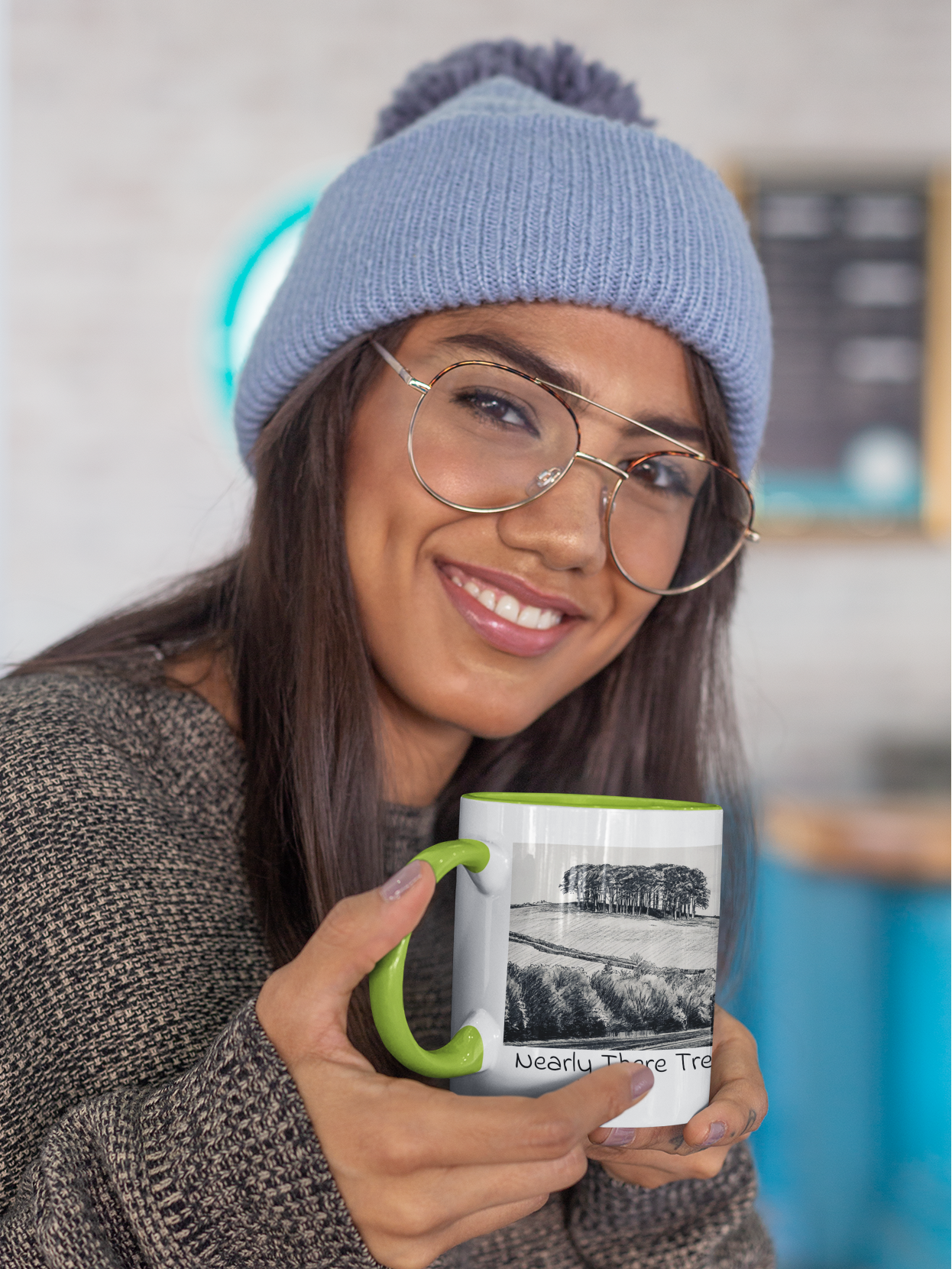 Woman in beanie holding Nearly There Trees mug with green handle, tree line pencil sketch design