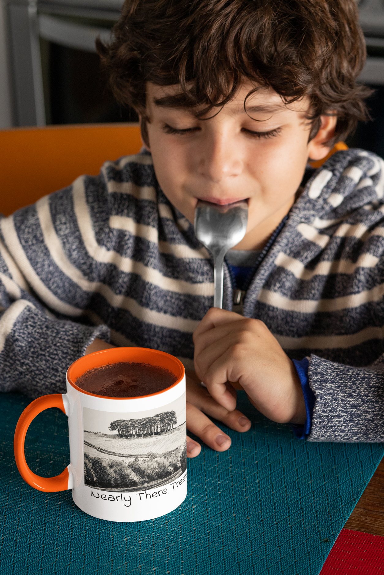 Boy with spoon beside Nearly There Trees mug with orange handle and hot chocolate, tree sketch design