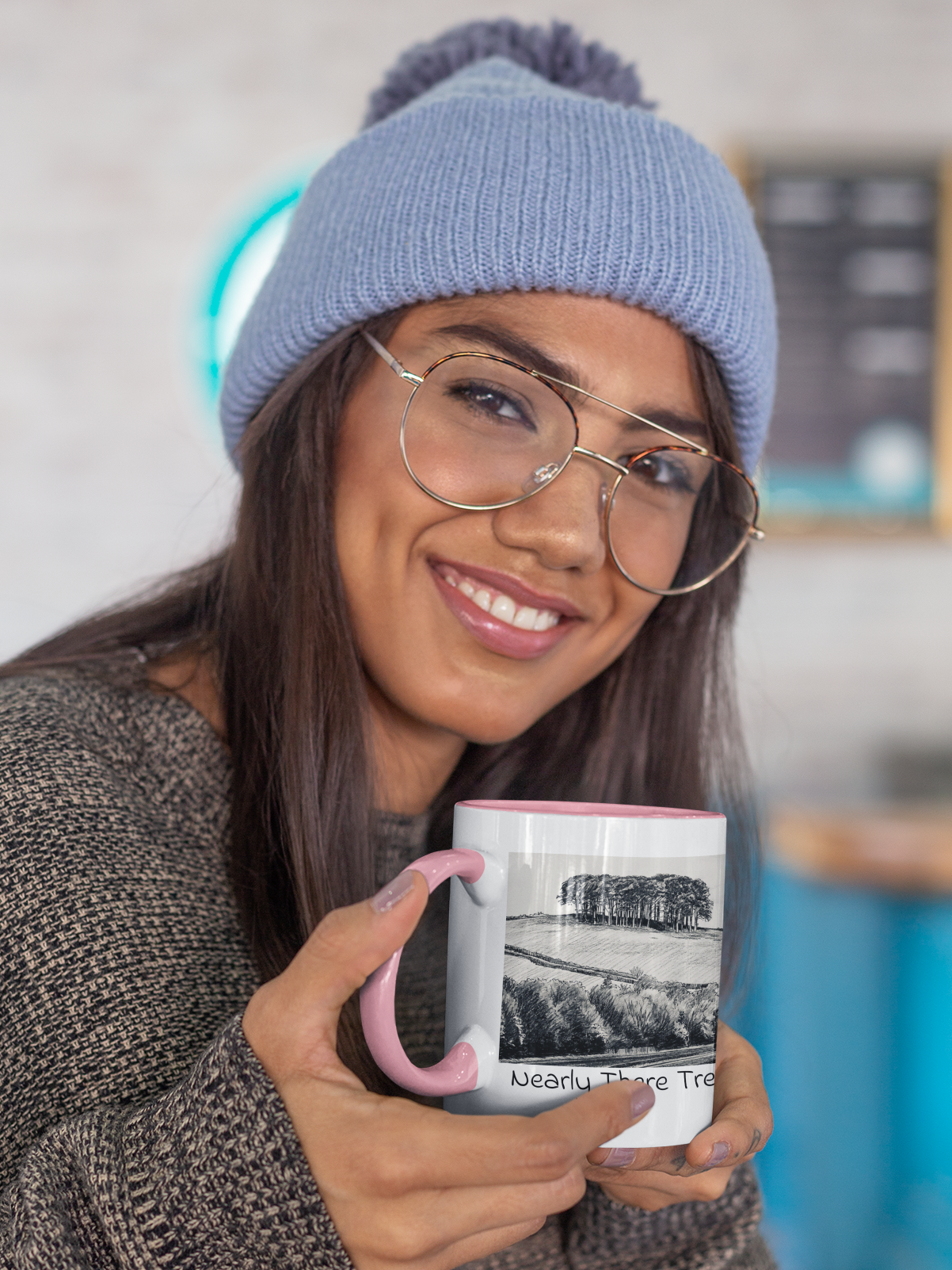 Woman in beanie holding Nearly There Trees mug with pink handle, tree line pencil sketch design